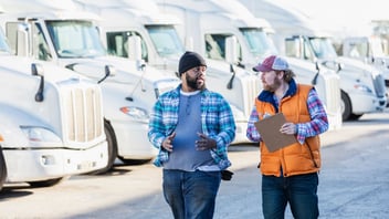 2 men talking, one holding a clipboard with a fleet of trucks in the background